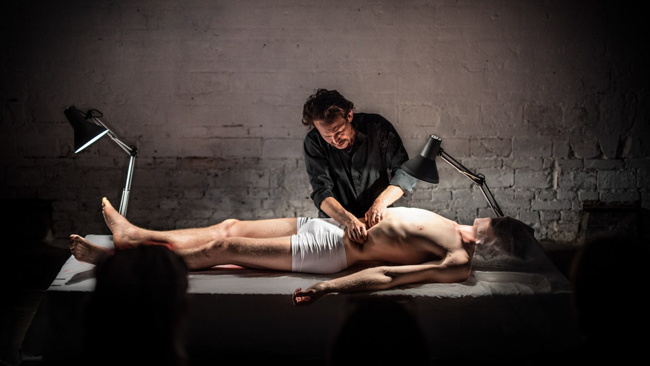 Photograph of a man lying on a table dressed only in underpants with a cloth wrapped over his face. Another man stands behind the table pressing on his stomach with his has. In the foreground are the backs of the heads of an audience.