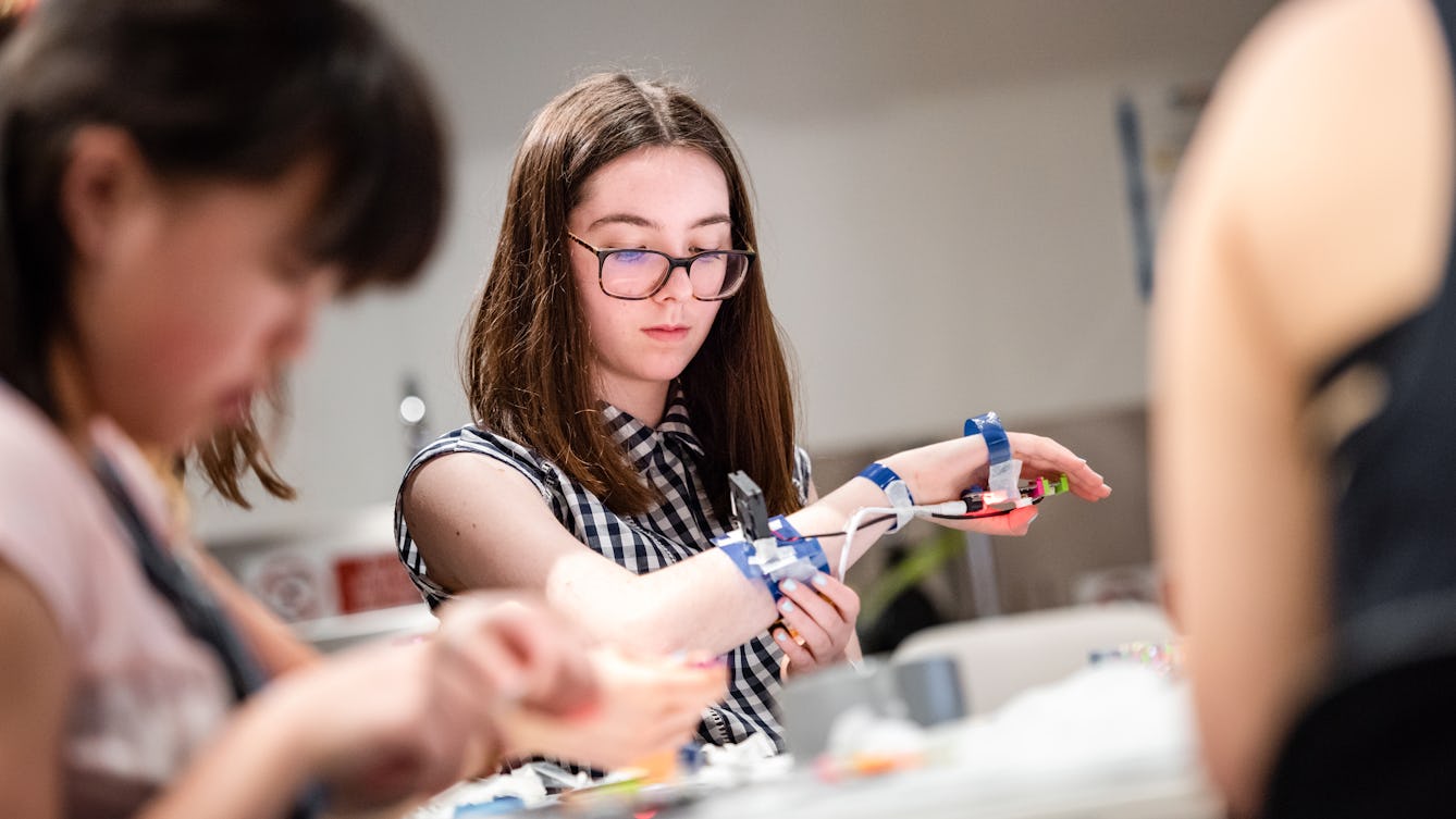 Photograph of a young girl taking part in a workshop to create a piece of technology you can wear. Her forearm has wires and components attached to it.