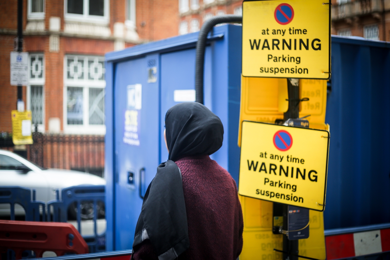 Photograph of a woman wearing a black head scarf facing away from the camera towards a street scene containing a blue building site storage container and two large yellow signs which say, "(no stopping symbol) at any time WARNING Parking suspension".