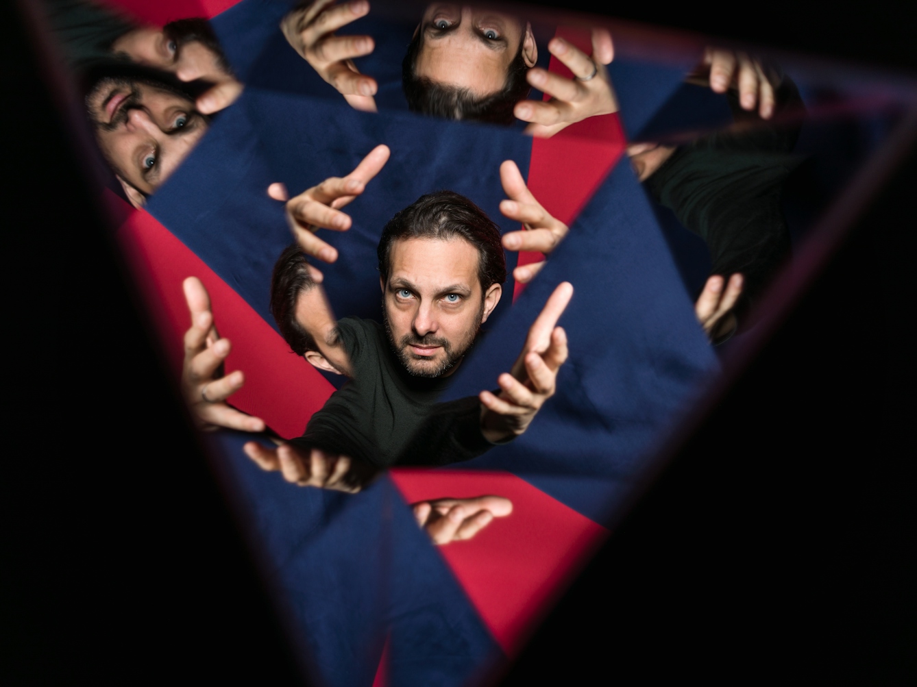 Photograph through a kaleidescope of the head of a man. His arms are stretched forwards, framing has face. The background is made up of blue and red fabric.