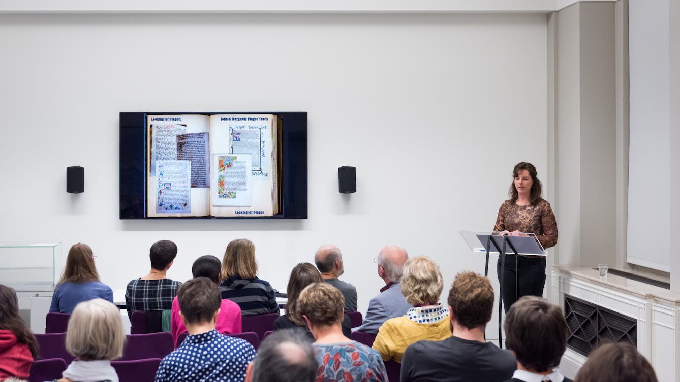 Photograph showing a woman giving a talk in the Viewing Room at Wellcome Collection. She is stood at the front of the room looking at a wall mounted television screen. In the foreground are the backs of the heads of the audience.