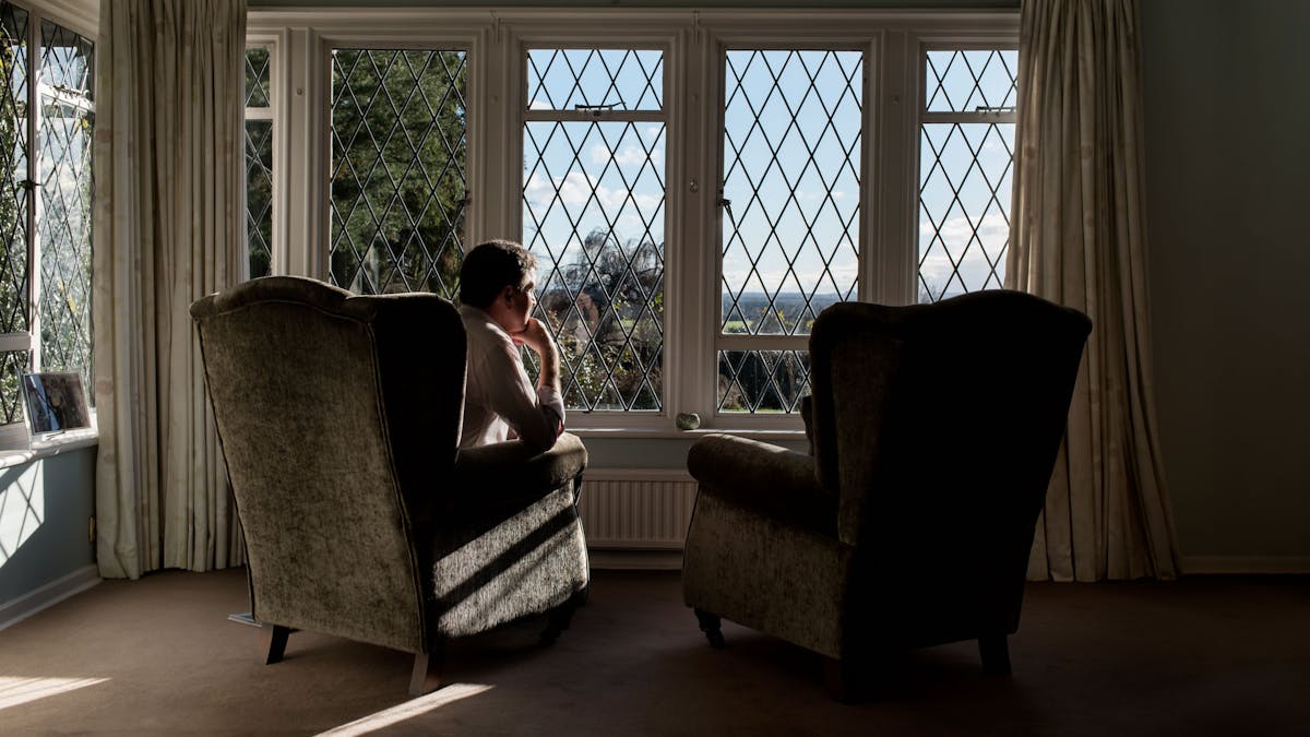 Photograph of a man sat in a living room armchair looking out of a window, his chin resting on his hand. To his right is an identical armchair which is empty,