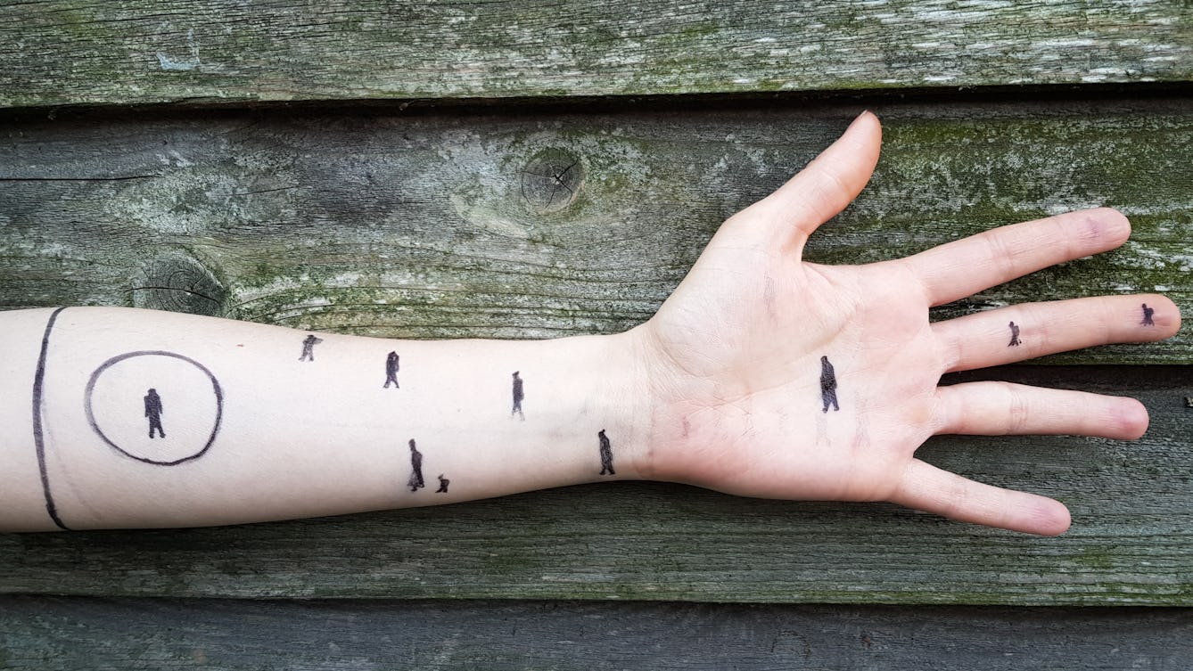 Photograph of a forearm with fingers outstretched, against a slatted wooden background. Small human figures are draw onto the arm, one is circled.