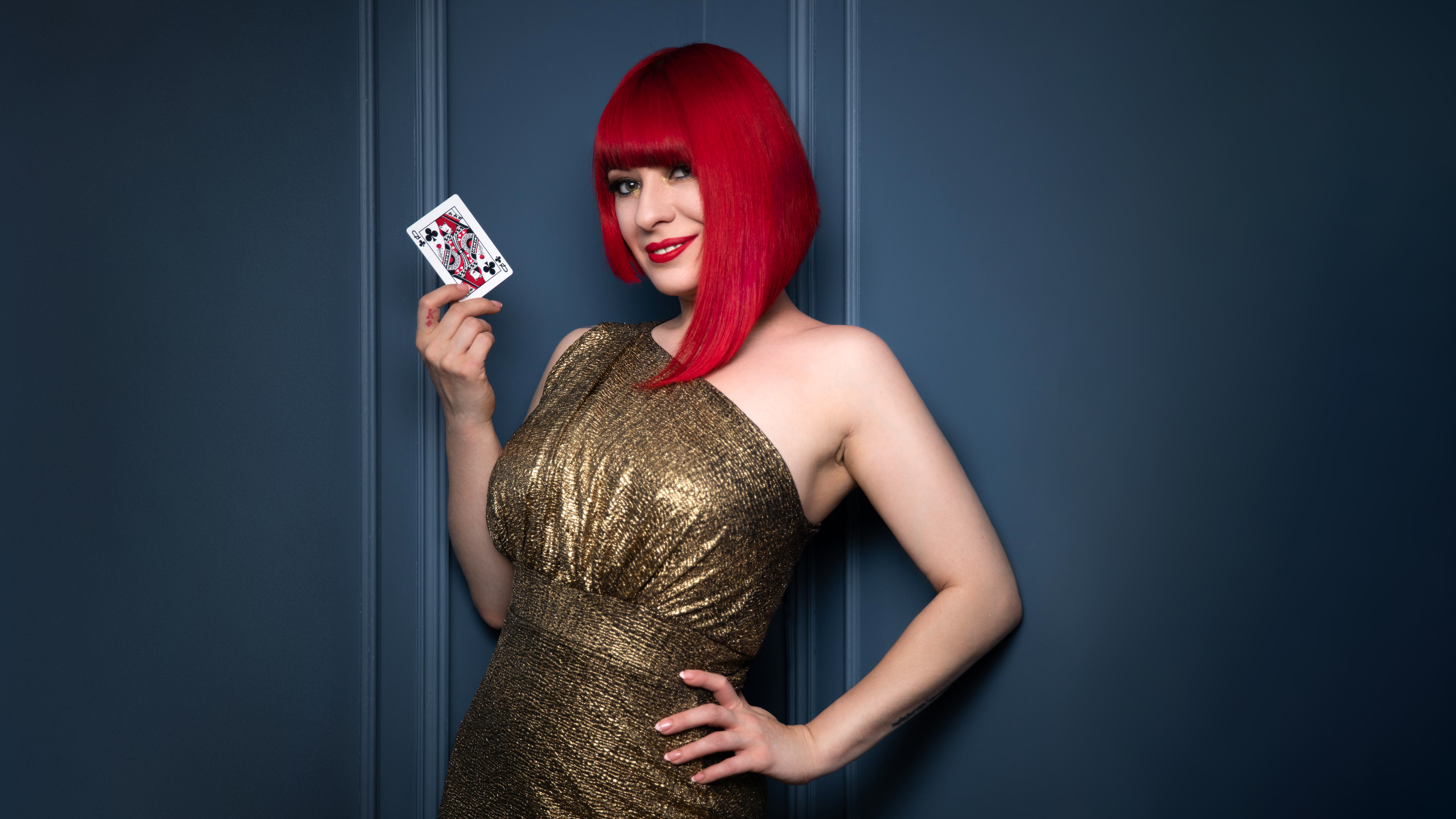 Photograph of a young woman with red hair, wearing a sparkling gold dress. She has one hand on her hip and the other is holding up the queen of spades playing card. Behind her is a blue wall.