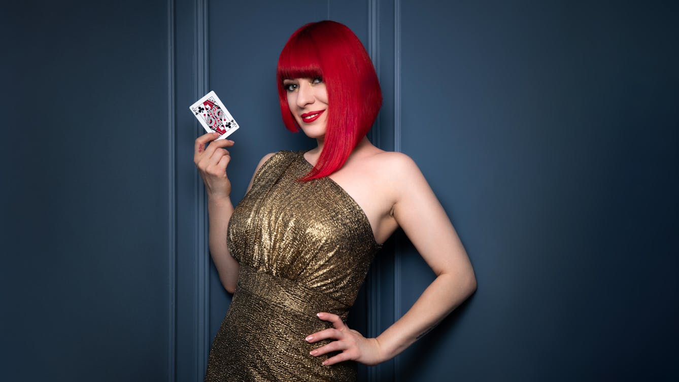 Photograph of a young woman with red hair, wearing a sparkling gold dress. She has one hand on her hip and the other is holding up the queen of spades playing card. Behind her is a blue wall.