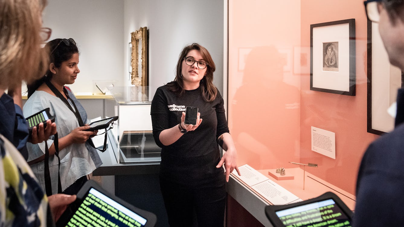 Photograph of a Visitor Experience Assistant conducting a speech to text tour of an exhibition at Wellcome Collection. She is talking into a phone whilst the visitors read a transcription of what she is saying from a tablet screen.