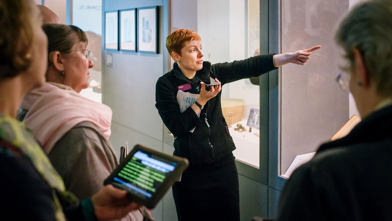 Photograph of a Wellcome Visitor Experience Assistant speaking into a phone whilst giving a speech-to-text tour of a gallery. In the foreground is a tablet displaying the transition.