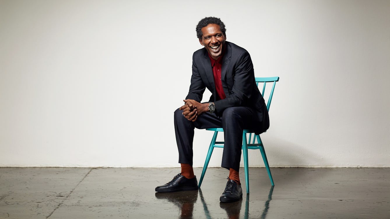 Photographic portrait of Lemn Sissay sitting on a wooden chair smiling.