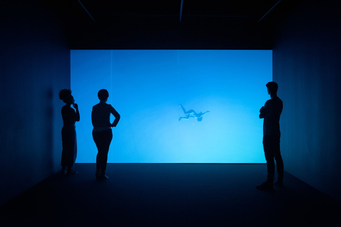 Photograph of visitors exploring a gallery installation. They are standing in the dark in front of a large projection of a figure floating underwater. The room has a blue cast.