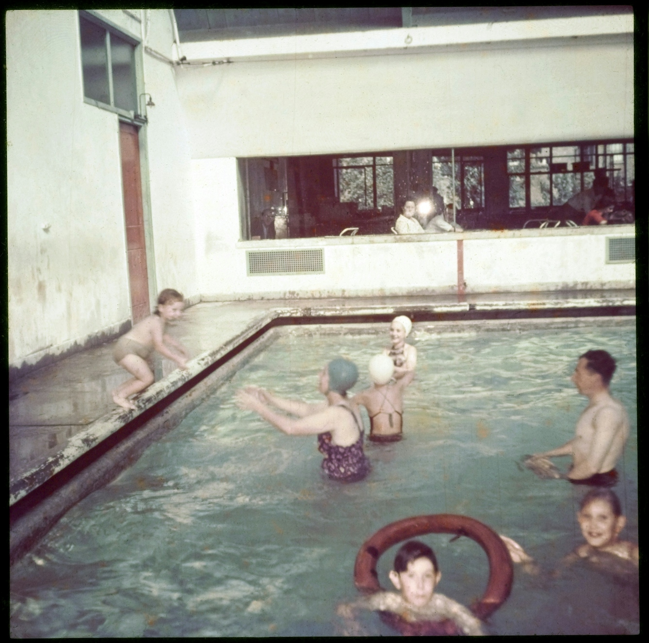Colour photograph of families in the pool at the Peckham Pioneer Health Centre, showing a child jumping in, a woman and girls wearing rubber swimming hats, and a child with an inflatable ring. In the background is a window through which people are sitting watching the swimmers.