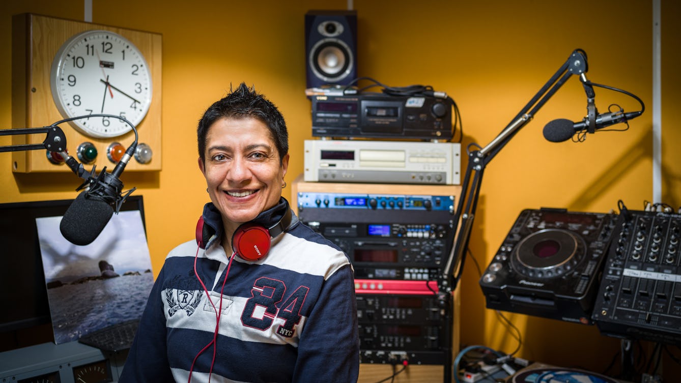 Photographic Portrait of a female DJ in a small Dj booth at a radio station. Behind the woman is radio equipment and a clock on the yellow sound absorbing wall.