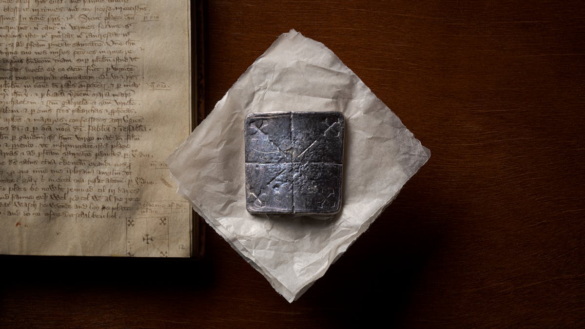 Photograph of a curved lead healing plate about ten centimetres square, sitting flat on a piece of conservation paper. Below the lead plate, to the left, is the manuscript from which the original instructions to make the plate can be found. The lead plate has been inscribed with 5 crosses; one diagonal cross in each corner with lines leading to a central, right-way-up, cross.