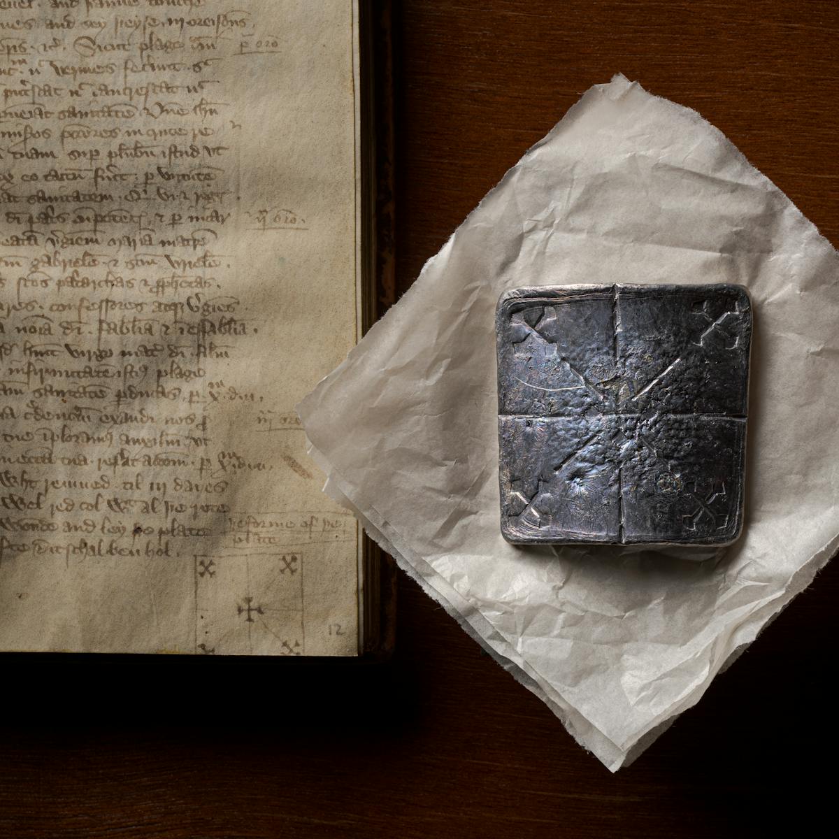 Photograph of a curved lead healing plate about ten centimetres square, sitting flat on a piece of conservation paper. Below the lead plate, to the left, is the manuscript from which the original instructions to make the plate can be found. The lead plate has been inscribed with 5 crosses; one diagonal cross in each corner with lines leading to a central, right-way-up, cross.