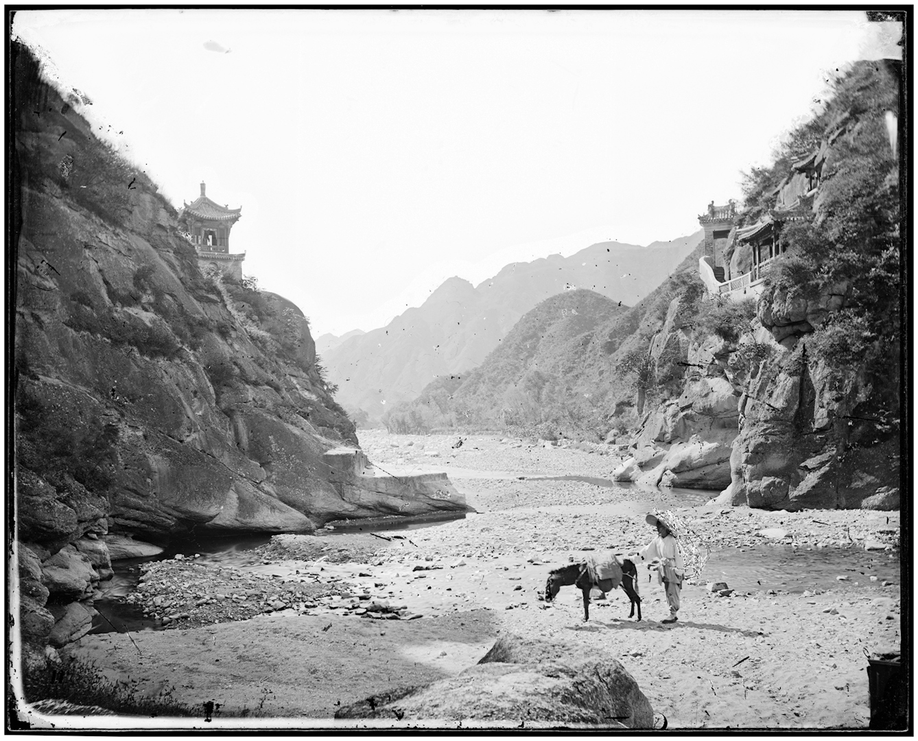 Black and white photograph of a mountain pass. A rough road runs between steep rocky cliffs, with wooden buildings balanced on them. A man and his donkey are in the foreground.