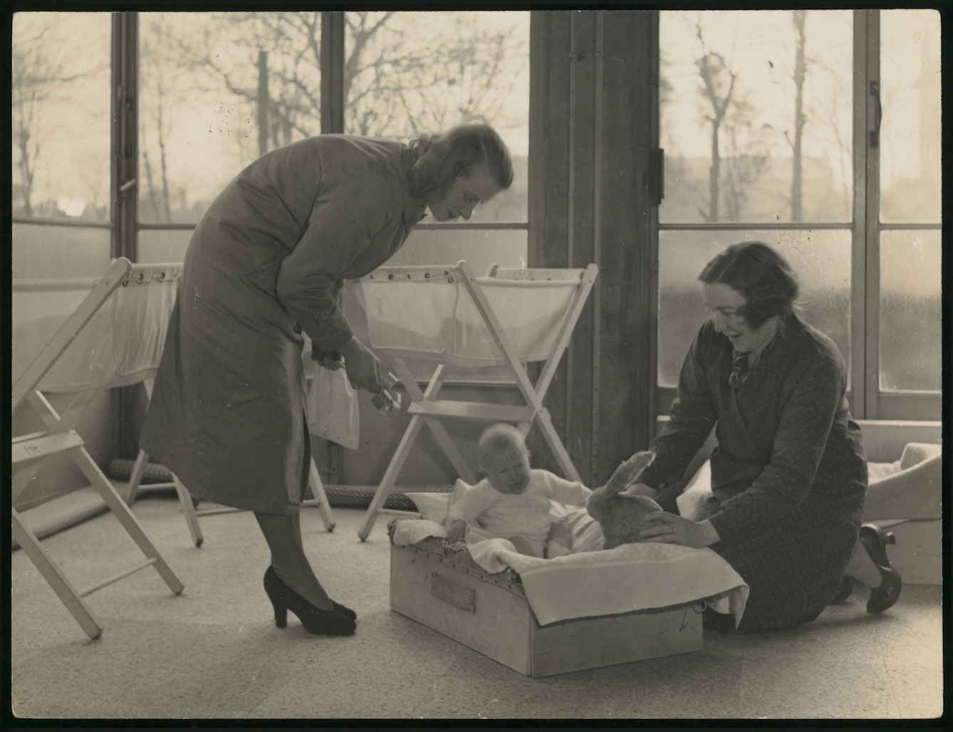Black and white photograph showing two women attending a baby in a cot.