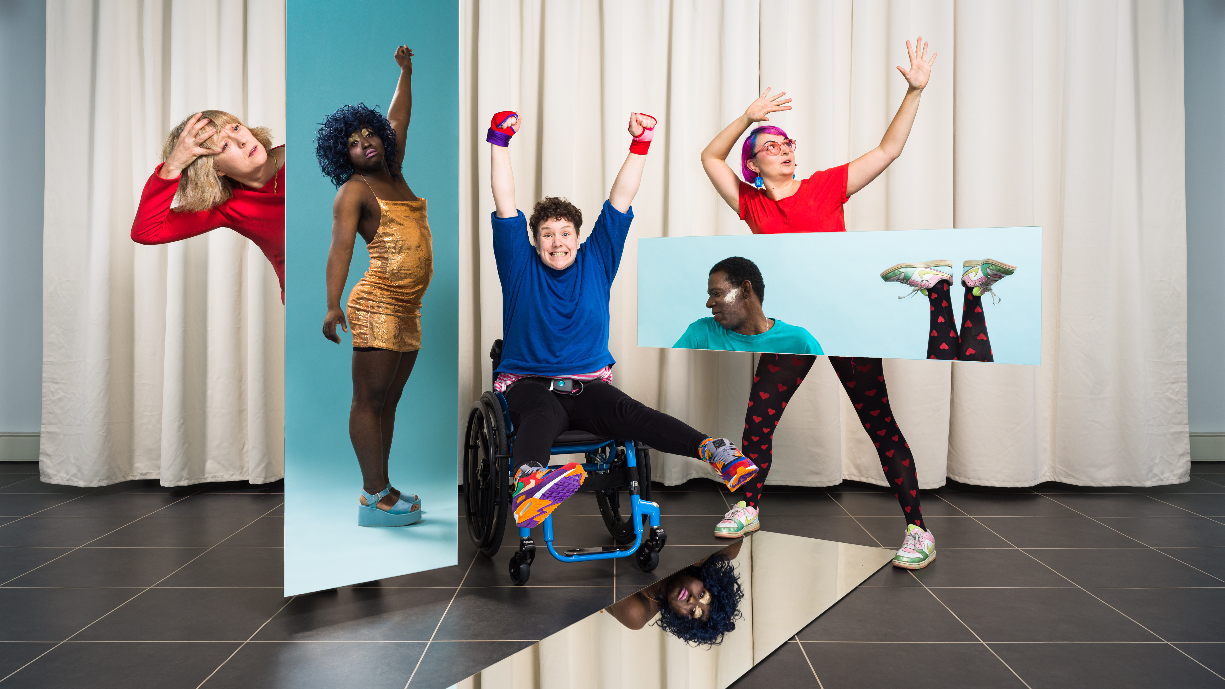A group of festival performers wearing colourful clothes, colourful make-up and striking poses. They are standing in front of cream curtains. Two mirrors are suspended in mid-air and another mirror is on the ground. The mirrors show reflections of the festival performers. 