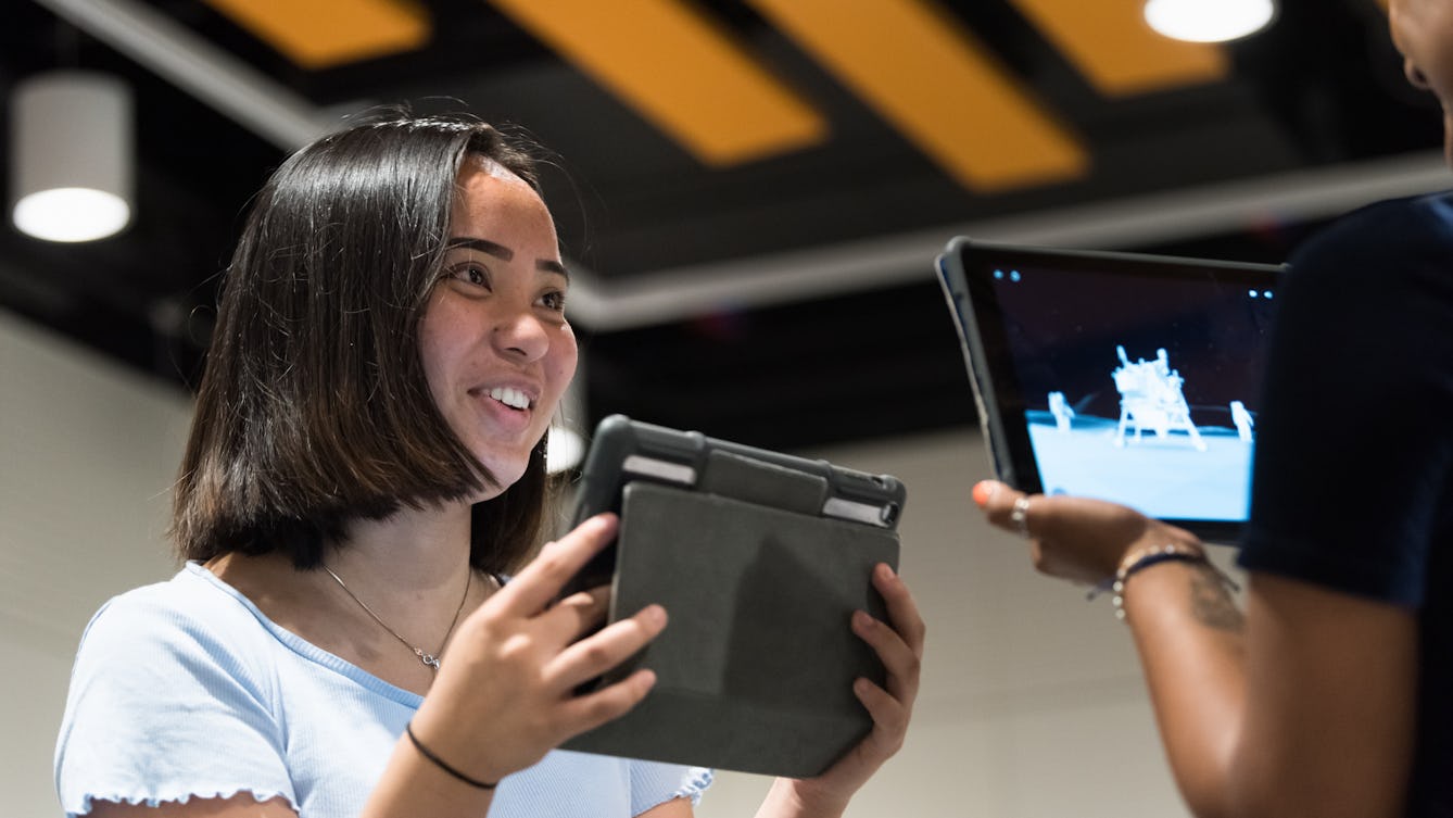 Photograph two young women standing in the Youth Studio at Wellcome Collection. Both young women are holding an iPad and smiling. On the iPad screen is an image of two astronauts on the moon. 