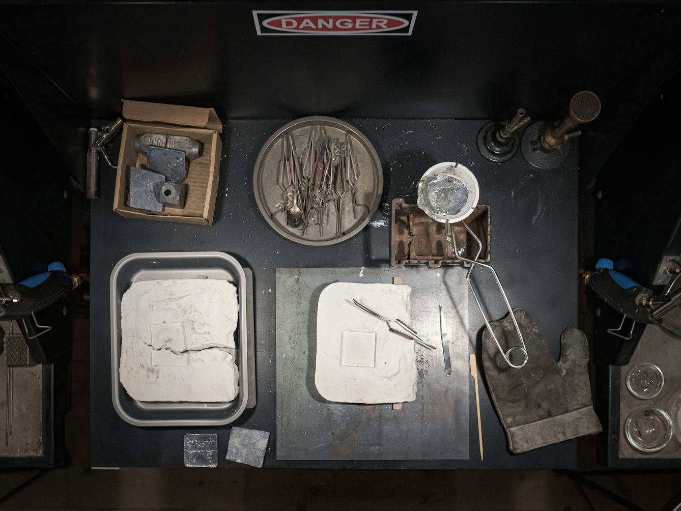 Photograph of instruments required to make the lead healing plate laid out on a workbench.  In the top left is a box containing raw lead.  In the centre us a dish of pliers, to the left is a ceramic bowl in the grip of a pair of tongs.  Inside the bowl is solidified lead, ready to be reheated.  to the bottom are two plaster-cast moulds, one of which is broken. The shape of the mould shows the imprint of a square.  In the bottom right there is a large heat-proof glove.  On the hood above the tools is a sign reading 'DANGER'.