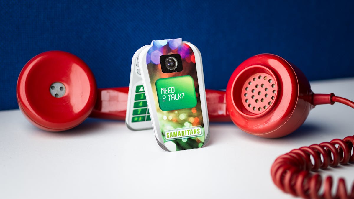 Photograph of an old red rotary phone receiver lying on its side on a white desk. Behind it is a blue felt desk back. between the earpiece and the mouthpiece is a Samaritans counseling service information leaflet in the shape of a flip mobile phone. on the front is written, 'Need 2 talk?'.