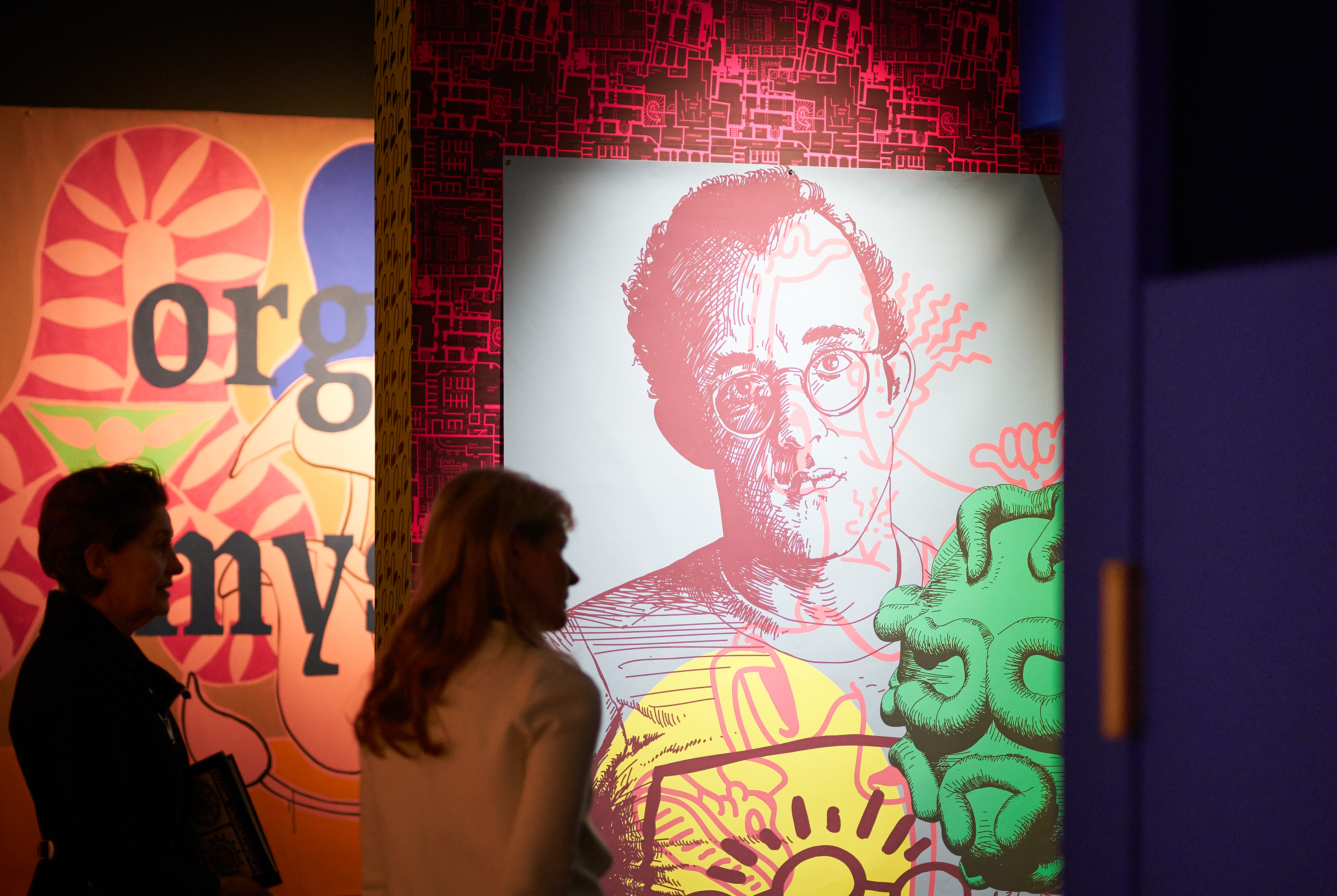 Photograph of visitors looking at colourful artworks displayed on coloured patterned wallpaper.