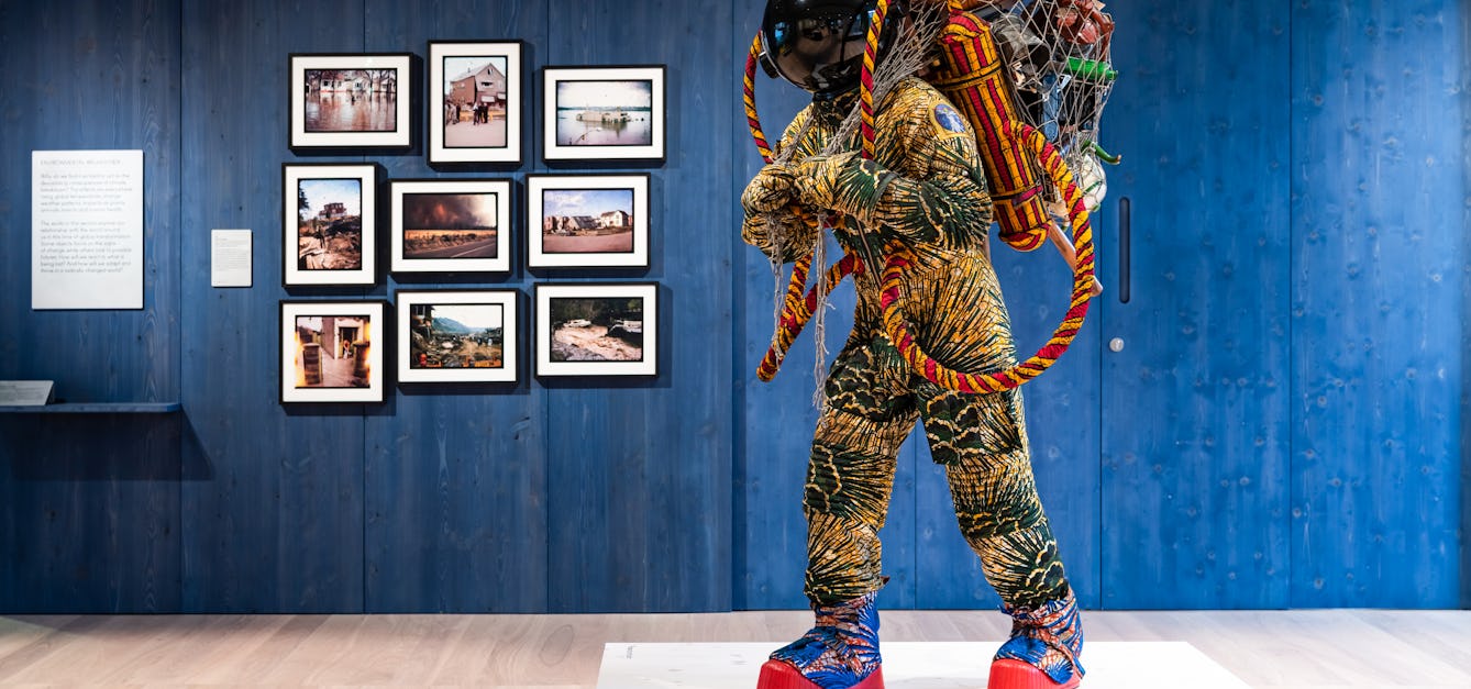 Photograph of an exhibition gallery space, with a blue stained wood wall in the background, on which are hung 9 photographs showing flooded landscapes. In the foreground is a life-size artwork of a figure resembling an astronaut. carrying a large net containing assorted objects including a suitcase.
