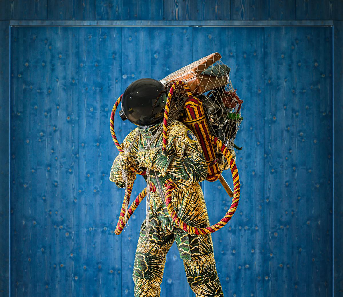 Photograph of an exhibition gallery space, with a blue stained wood wall in the background. In the foreground is a life-size artwork of a figure resembling an astronaut. carrying a large net containing assorted objects including a suitcase.
