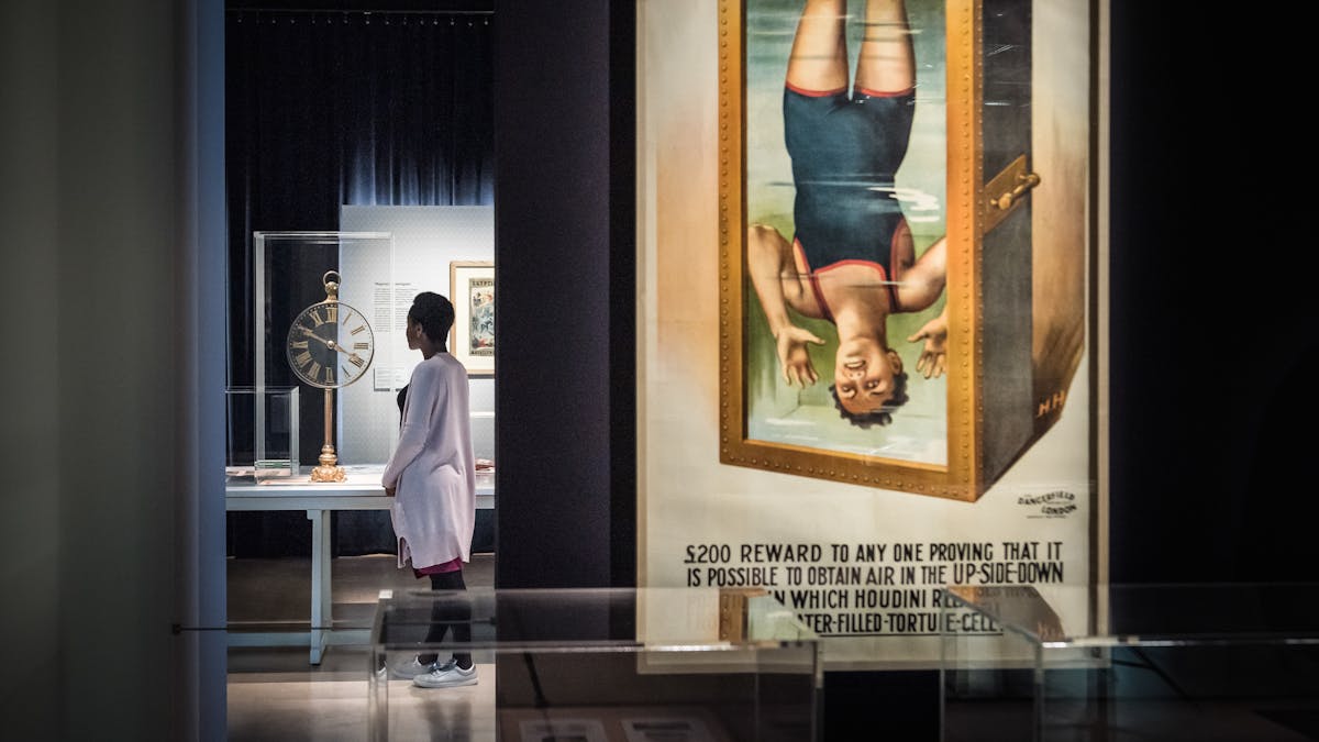 Photograph of a visitor exploring the exhibition, Smoke and Mirrors at Wellcome Collection. Photograph shows a women in the background looking at an object in a display case. In the foreground is a large colourful poster showing a man in a Victorian swimming costume suspended upside down in a tank of water.