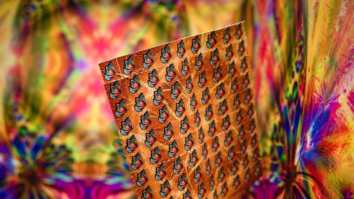 Photograph of a pretend sheet of LSD tabs with a butterfly motif, against a colourful abstract background.