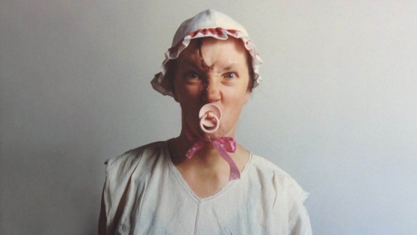 Photographic self-portrait, from the chest up, of Jo Spence as a grown woman, wearing a babies bonnet and sucking a dummy.