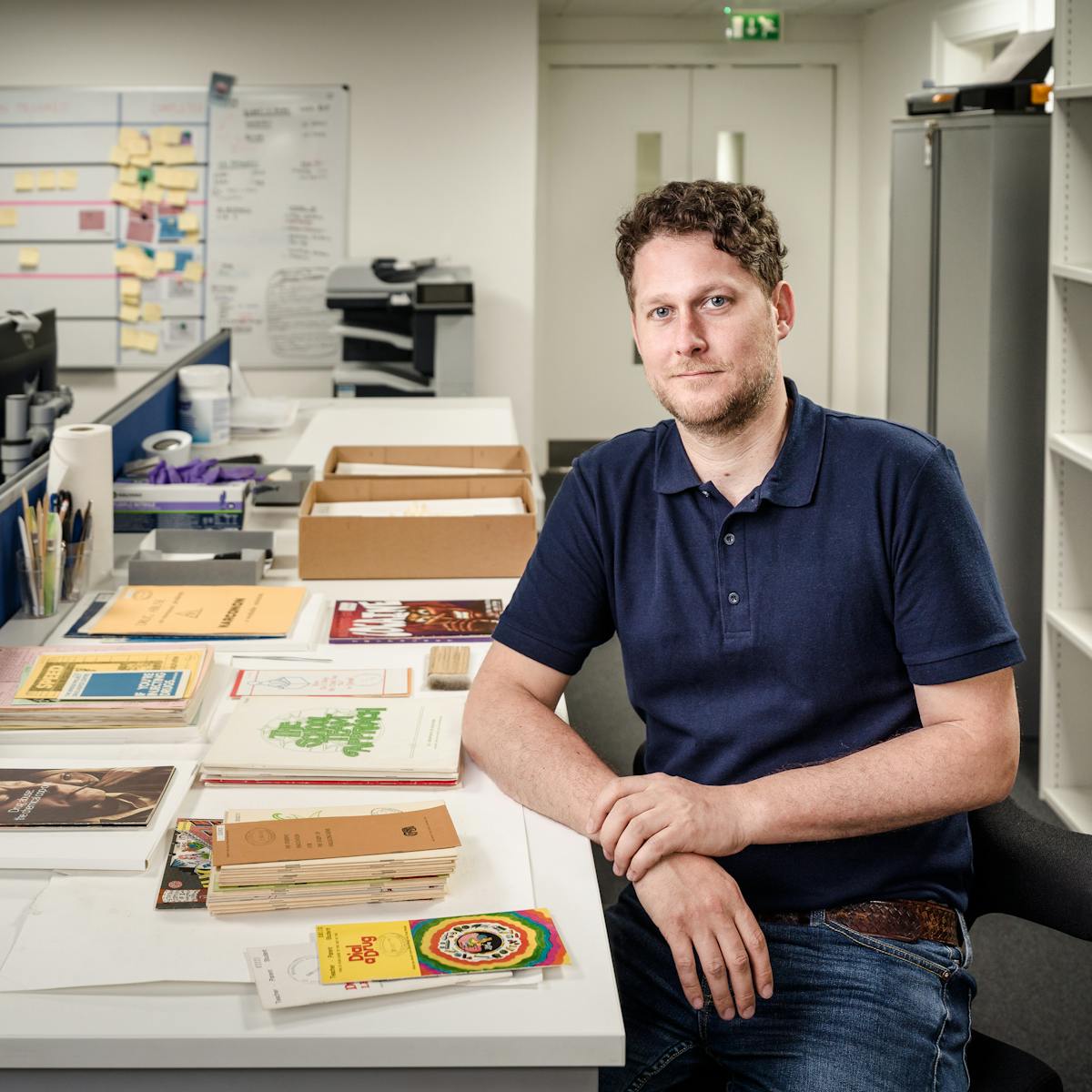 Photograph of a man sitting at a long desk in a library archive preparation room. On the table in front of him is various leaflets and books from the archive. To his left are shelves containing books.