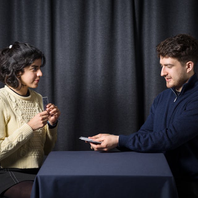 Photograph of a young man and woman performing magic tricks with a deck of playing cards, in front of a grey curtain.