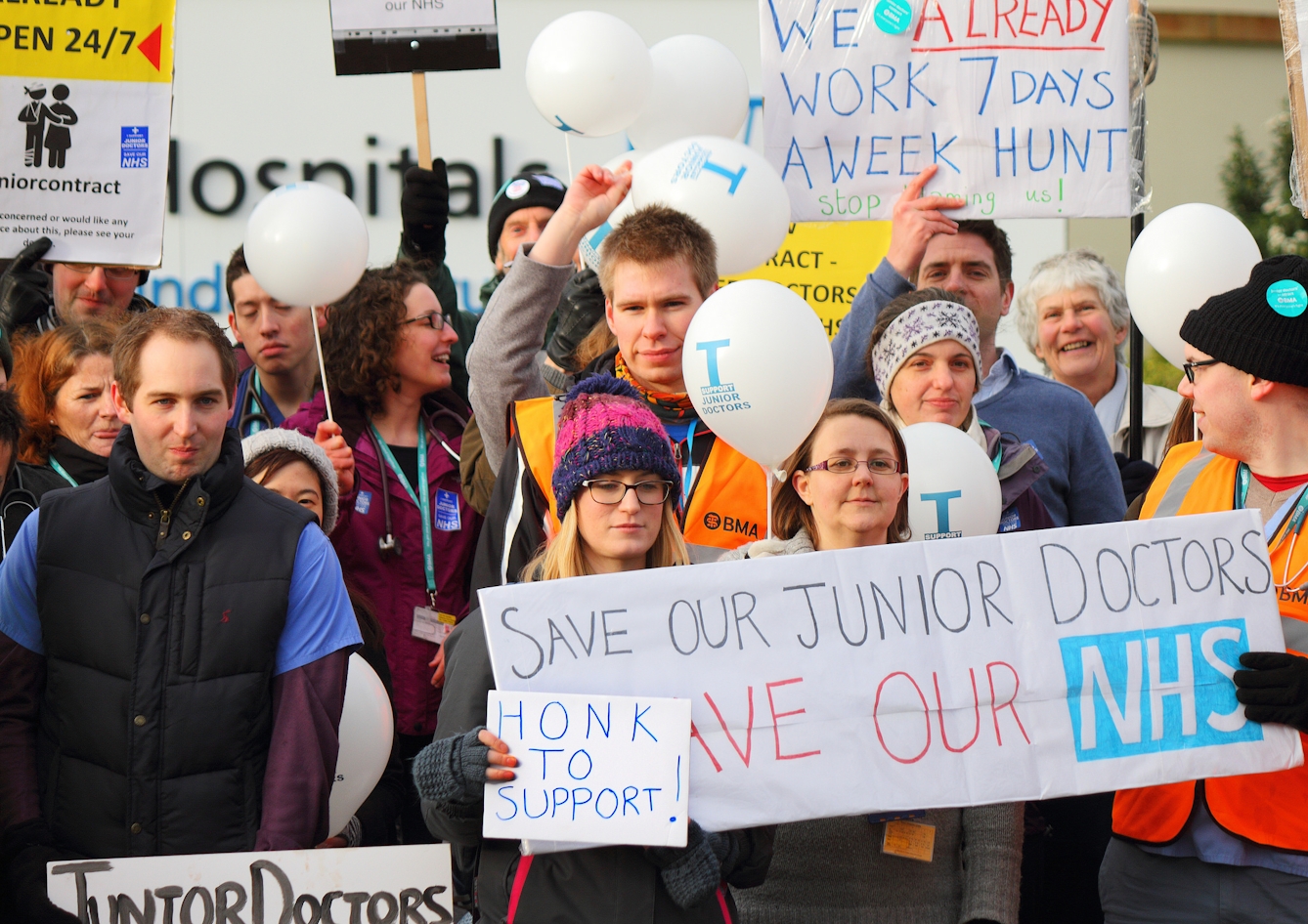 Colour photograph of a group of people on strike. They wear coats, gloves and hats, and some wear high-visibility jackets that have "BMA" printed on them. Placards and banners include "We already work 7 days a week Hunt" and "Save our junior doctors. Save our NHS."