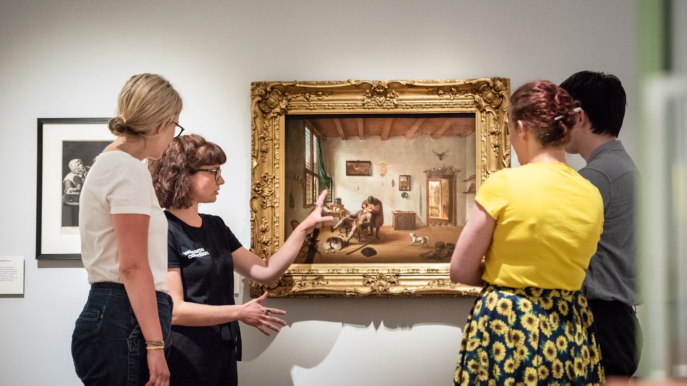 Photograph showing a visitor experience assistant, standing in front of an oil painting, giving a guided tour of an exhibition to three visitors.