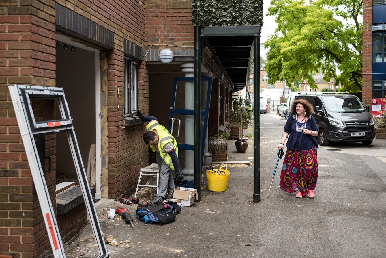 Photograph of a woman walking with a crutch down an alley, past a workman installing windows in a property.