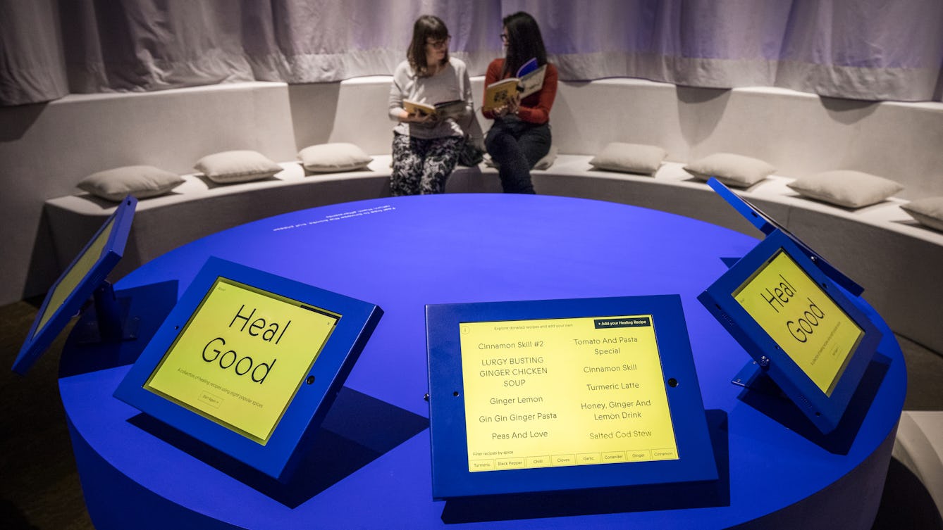 Photograph of two out-of-focus women discussing a book in the background. In the foreground are five exhibition tablet devices with interactive content. One has the title text 'Heal Good' displayed, another has a list of ingredients.