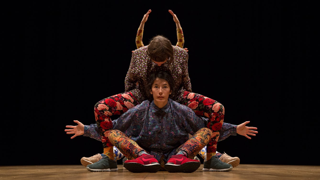 Dancers wearing colourful floral clothing creating shapes as they move together. Performing in a studio with a black backdrop and wooden floors. 