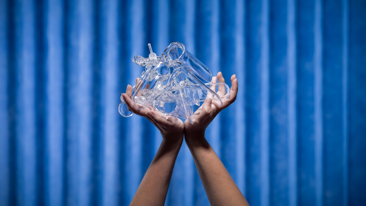Photograph of two hands struggling to hold several vaginal speculums shot against the background of blue antibacterial clinical curtains.