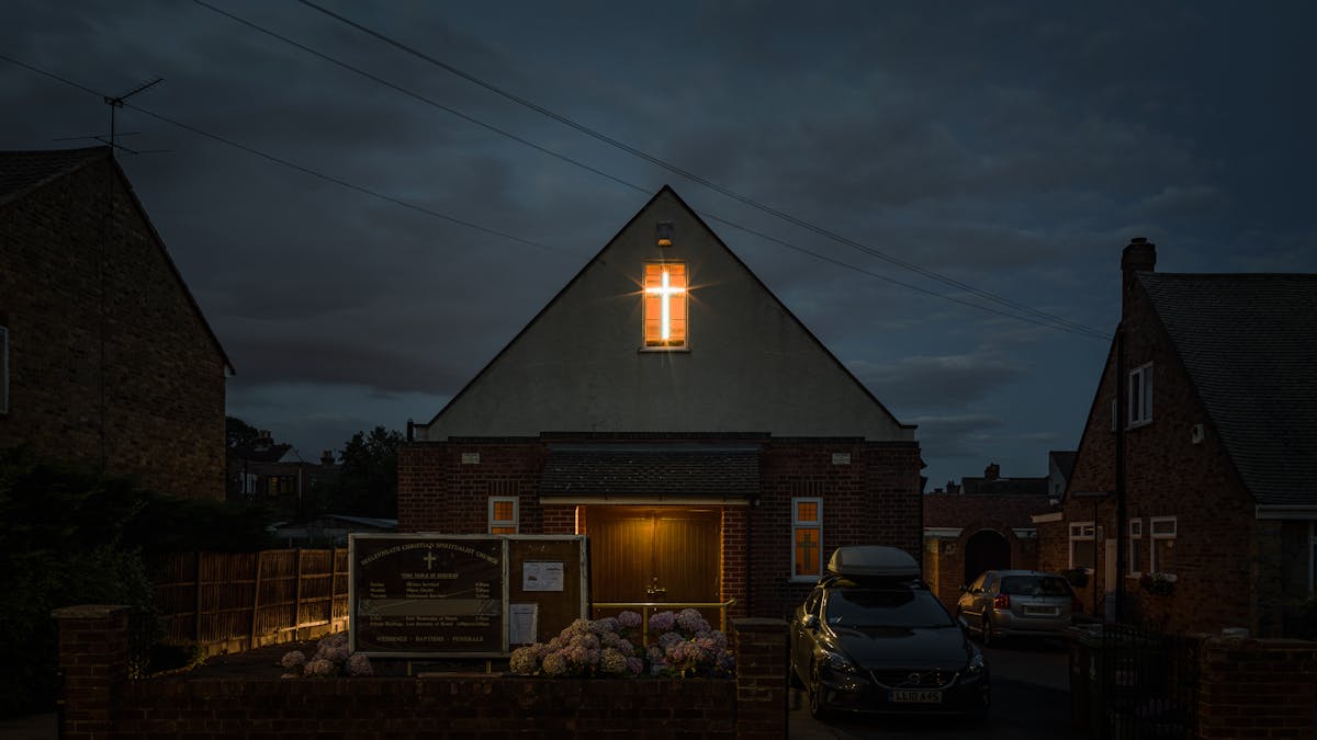 Photograph of Bexleyheath Christian Spiritualist Church at night. The two storey building is set in a residential street, detached but sandwiched between other residential buildings. The second storey shows a brightly lit window with a cross shaped light showing in the window.  To the right of the building are two cars.