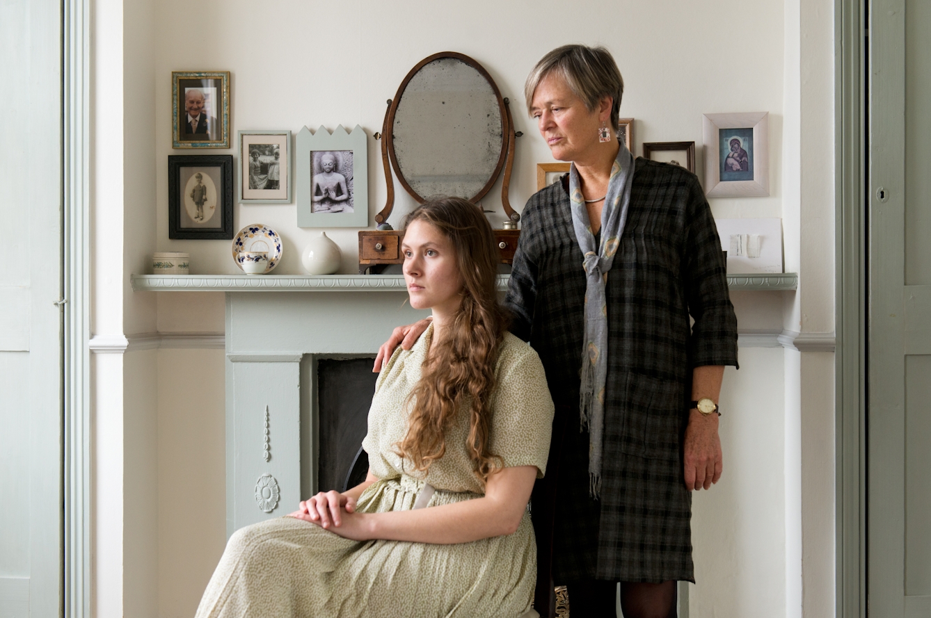 Photograph of a young woman and a mother figure in a domestic living room. The young woman is seated in a long dress and and the mother figure stands behind her, hand on her shoulder. They are both gazing out of the frame.