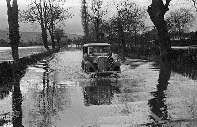 A black and white photograph showing a 1930s model car driving through floodwaters.