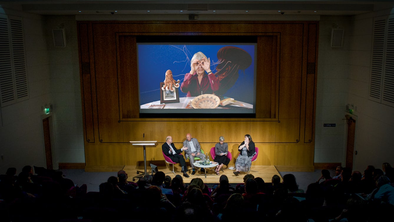 Photograph of a dark auditorium showing the backs of the audience's heads and four panel members seated on a small stage, in-front of a large screen. On the screen is a still from a film.