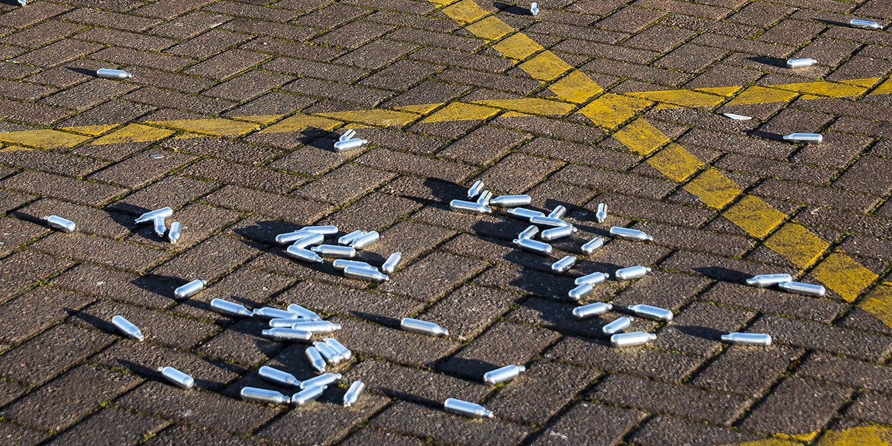 Image of numerous small silvery cannisters known as "whippets" that contain nitrous oxide gas. The whippets are scattered across the ground in a car park, indicated by painted lines.