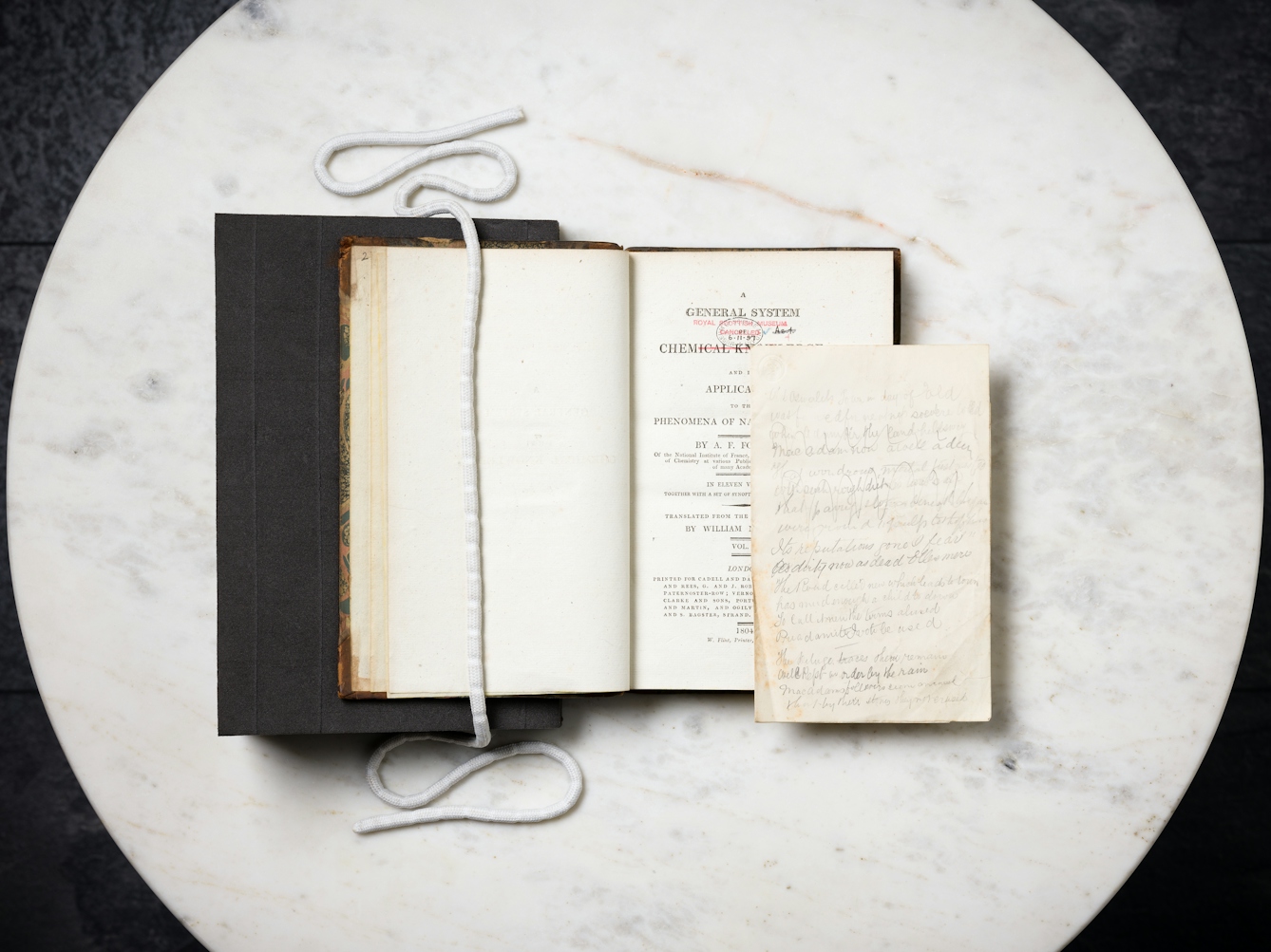An old book propped open on a marble tabletop, with a piece of paper bearing pencilled verse resting inside.