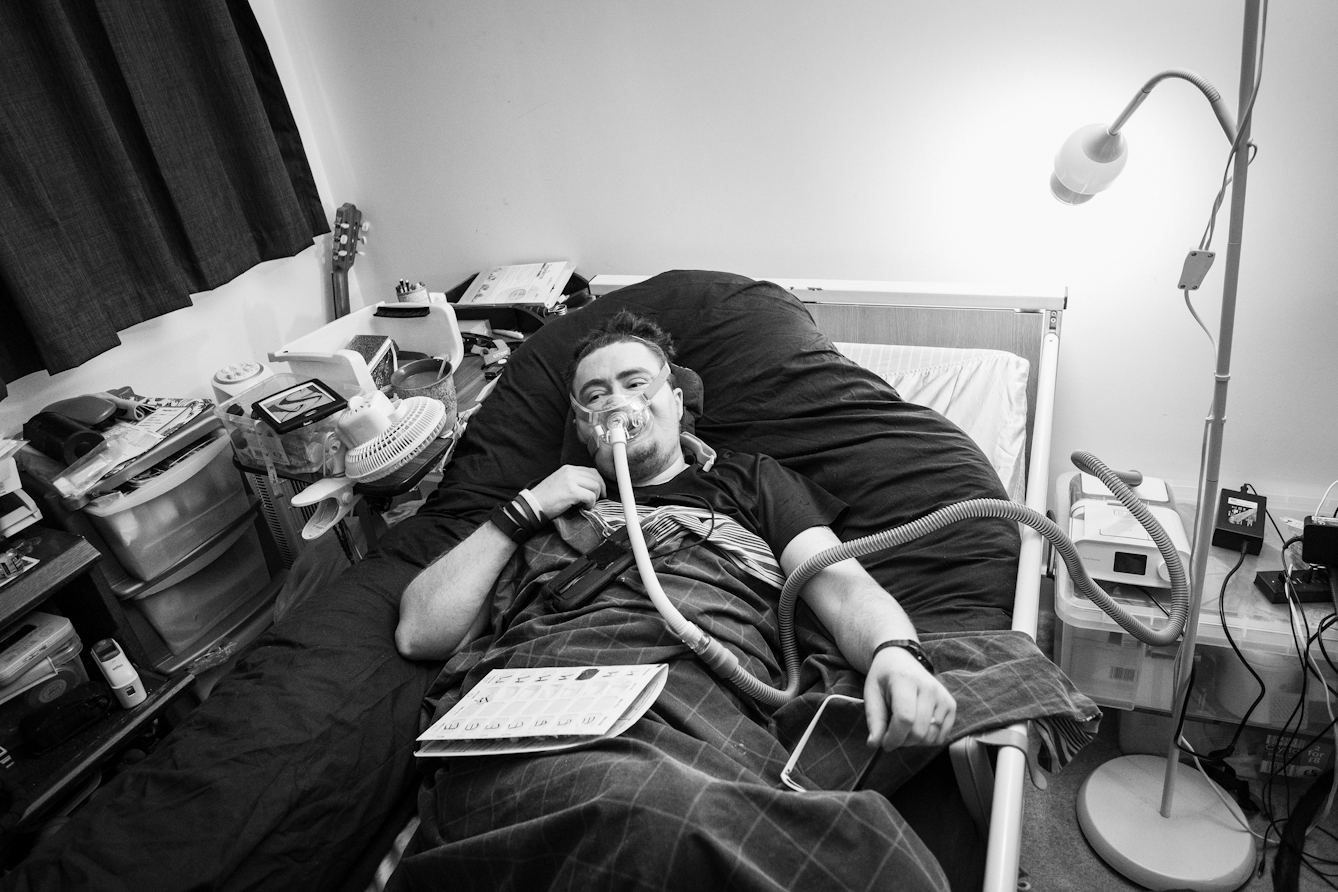 Black and white photograph of an individual at home in bed using a ventilator.