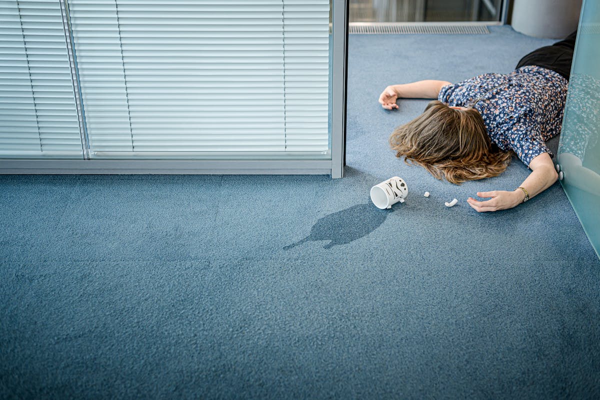 Photograph of woman lying face down on the blue carpet of an office space. Her arms a outstretched beside her head and in front of her on the floor is a white mug surrounded by the broken pieces of the handle. The liquid contents of the mug is spilt on the floor. 