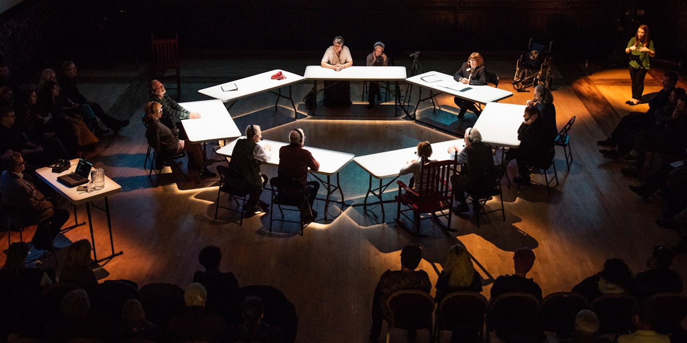 Photograph of a ring of spot lit tables at which people are sitting, in discussion. Around the outside of the circle is an audience and sign language interpreter. 