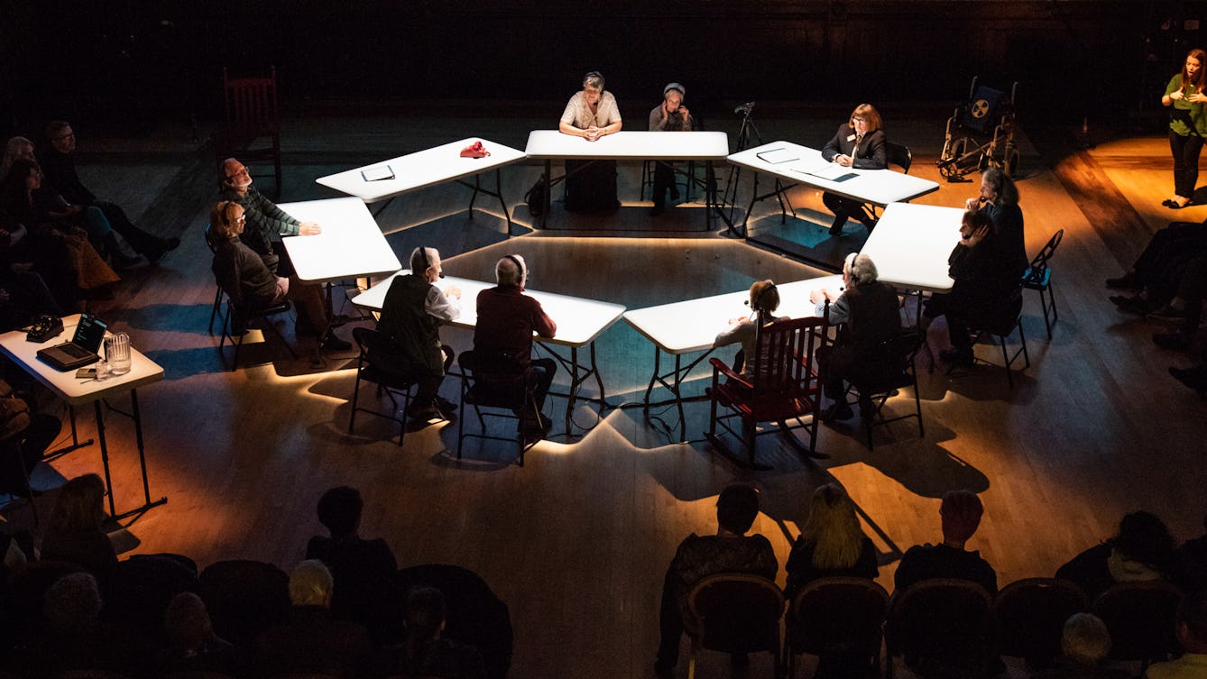 Photograph of a ring of spot lit tables at which people are sitting, in discussion. Around the outside of the circle is an audience and sign language interpreter. 