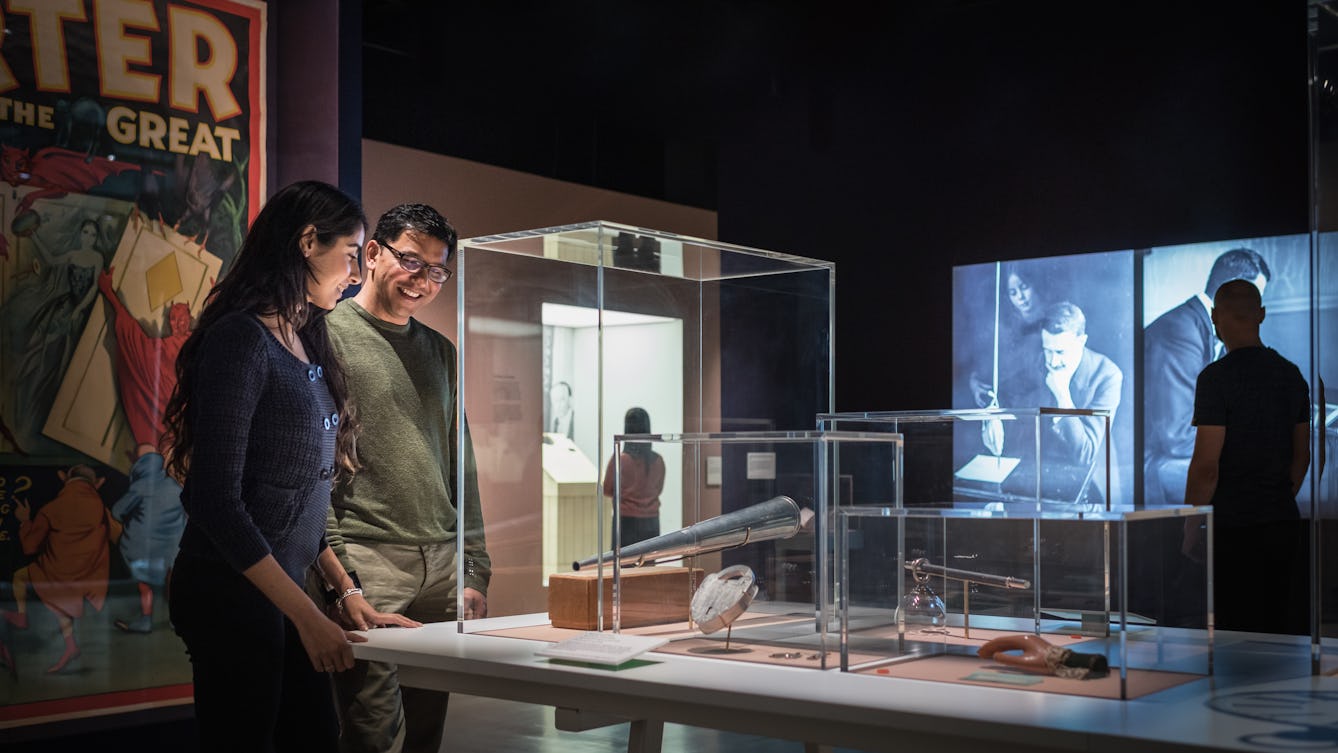 Photograph of visitors exploring the exhibition, Smoke and Mirrors at Wellcome Collection. Photograph shows a man and a women in the foreground discussing an object in a display case. Behind them is a large colourful poster and other gallery visitors.