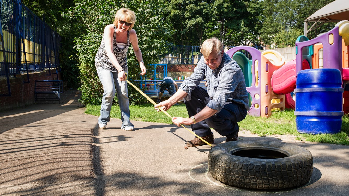Photograph of two adults in a children's playground. The woman is standing and smiling holding a tape measure. The man in the photograph is crouching near a car tyre on the ground. 