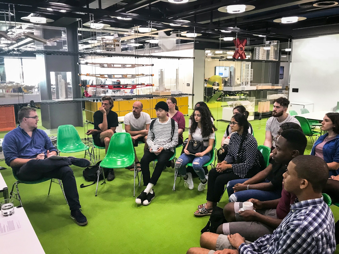 Photograph of a group of young people sitting in a semi-circle at an architects firm, listening to a man speaking.